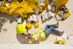 A construction worker lying unconscious on the ground with a colleague checking on them.