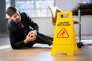 A man in a suit has fallen on the floor. He is clutching his knee in pain. There is a yellow wet floor sign next to him.
