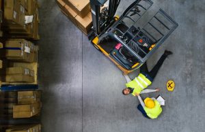 A man checking on his colleague on the floor who has been hit by a forklift in a warehouse.