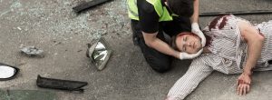 A person lying on the ground unresponsive with blood pouring from their head being attended to by a first aid trained person after a fatal accident. 